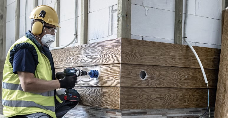 Handwerker mit Schutzkleidung bohrt Loch in Holzverkleidung mit Akkubohrmaschine